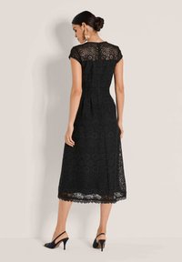 Schwarzes Spitzenkleid mit kurzen Ärmeln, floralen Ausschnitten und einem Oberschenkel langen Saum. Besitzt eine taillierte Taille und einen gewellten Saum. Tragen mit Riemchen-Heels.