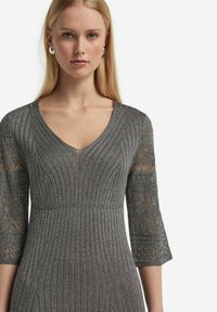 Grå stickad klänning med V-ringning, ribbad kropp och spetsärmar i trekvarts längd med geometriska mönster, tillverkad av ett strukturerat metalliskt material.