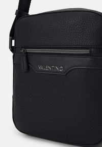 Black textured leather crossbody bag featuring a front zip pocket and subtle branding, with a smooth strap for adjustable carrying.