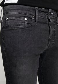 Primer plano de la cintura y la parte superior del muslo de unos vaqueros negros, mostrando los pasadores del cinturón, el bolsillo frontal y los detalles del cierre de botón.