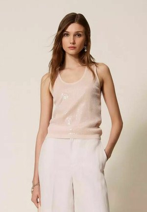 Giovane donna con lunghi capelli castani, indossa una canotta rosa chiaro senza maniche con paillettes e pantaloni bianchi a vita alta, in piedi davanti a uno sfondo semplice.