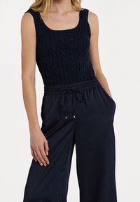 Kvinna som bär en ärmlös marinblå kabelstickad topp och marinblå vida byxor med dragsko, med händerna i fickorna.