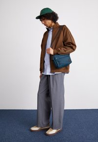 Giacca marrone con zip, camicia a righe celesti, pantaloni grigi a gamba larga, borsa a tracolla turchese e sneakers beige con suole marroni. Cappello verde.