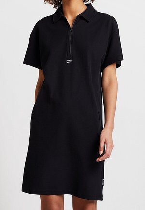 Frau trägt ein kurzärmeliges schwarzes Polokleid mit einem Reißverschluss vorne und dem Puma-Logo auf der Brust, steht vor einem einfarbigen Hintergrund.