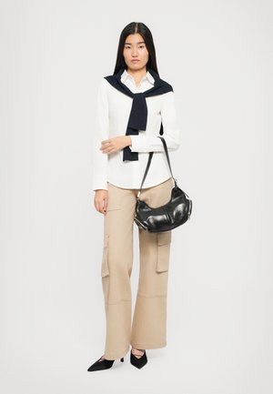 Woman wearing a white shirt, beige wide-leg pants, black heels, navy sweater draped over shoulders, holding a black leather handbag on her arm.