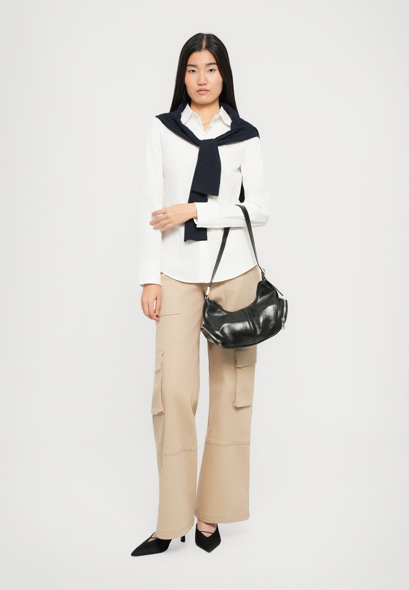 Donna in camicia bianca, pantaloni beige a gamba larga, tacchi neri, maglione blu navy drappeggiato sulle spalle, tiene una borsa in pelle nera sul braccio.