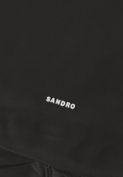 Tessuto nero con texture caratterizzato da un piccolo logo bianco "SANDRO". La superficie presenta sottili righe, con dettagli di cucitura visibili ai bordi.
