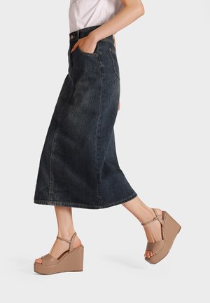 Gonna maxi in denim blu scuro con tasche laterali, abbinata a sandali con zeppa color beige dotati di cinturini alla caviglia e una finitura liscia.