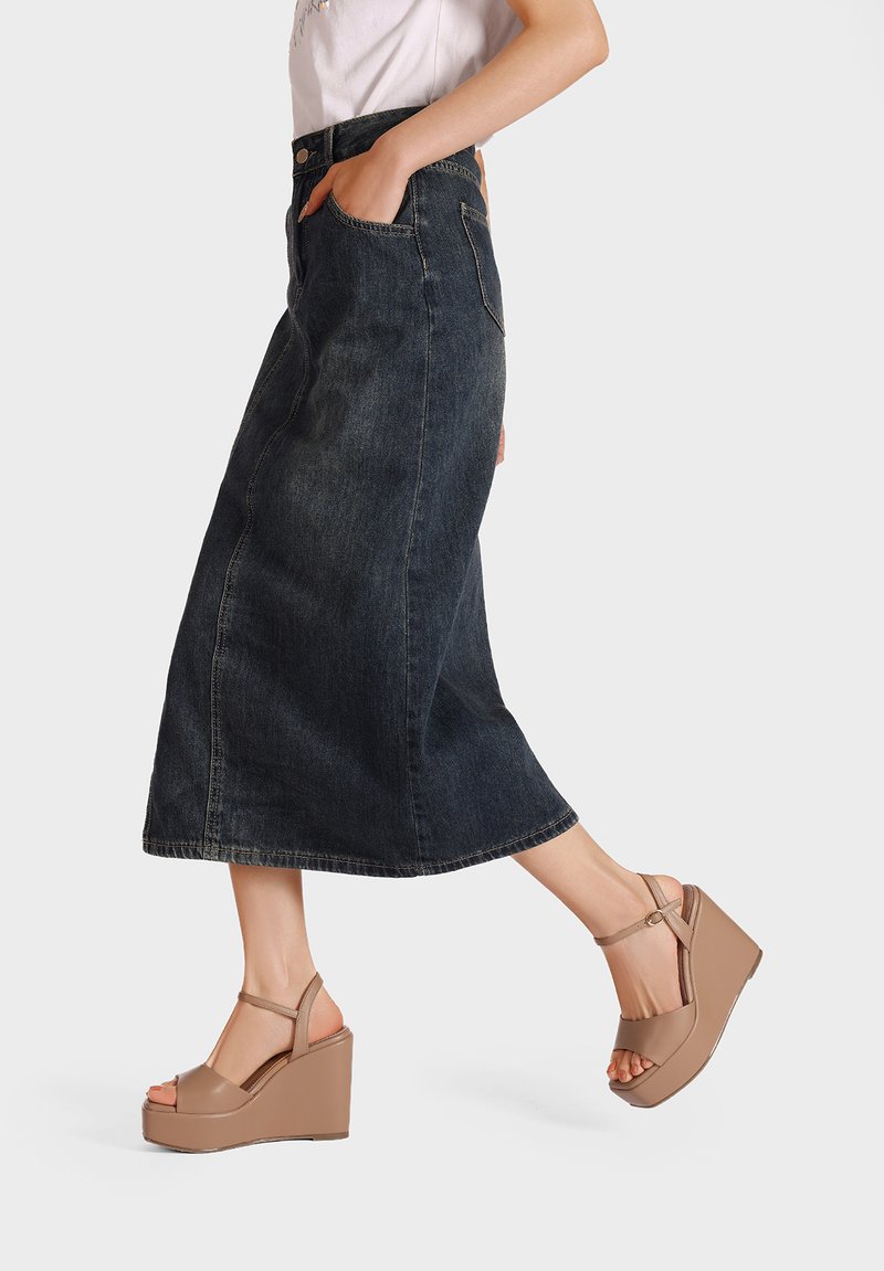 Gonna maxi in denim blu scuro con tasche laterali, abbinata a sandali con zeppa color beige dotati di cinturini alla caviglia e una finitura liscia.