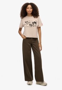 Jonge vrouw met krullend haar die een beige T-shirt met opdruk, bruine wijde broekspijpen en beige sneakers draagt, staand met één hand in haar zak.