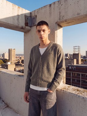 Giovane uomo che indossa un cardigan grigio e una camicia bianca si trova sul tetto con panorama urbano e cielo sereno sullo sfondo.