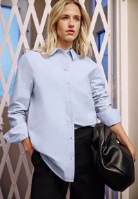 Vaaleansininen pitkähihaisen kauluspaita, yhdistettynä mustiin housuihin ja teksturoidun mustan laukun kanssa. Paita on väljä ja pehmeästä kankaasta.