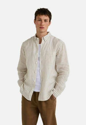 Jeune homme debout les mains dans les poches, portant une chemise à rayures beige et blanche boutonnée par-dessus un t-shirt blanc et un pantalon marron.