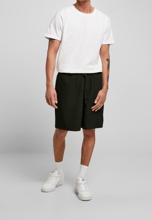 Weißes T-Shirt mit kurzen Ärmeln und schwarze elastische Shorts. Das Shirt hat eine einfarbige Gestaltung; die Shorts haben einen Kordelzug und einen lässigen Schnitt. Weiße Sneaker runden den Look ab.