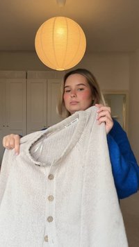 Beżowy sweter z dzianiny z okrągłym dekoltem, z pięcioma drewnianymi guzikami i widocznym teksturowanym ściegiem. W tle znajduje się duża lampa wisząca.