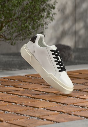Baskets blanches avec lacets noirs et détail au talon flottant au-dessus d'une surface en blocs de bois en extérieur, près d'un feuillage vert et d'un mur en béton.