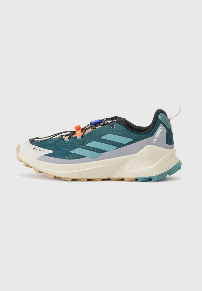 Chaussure de randonnée Adidas Terrex bleue et grise avec semelle beige, lacets noirs et fermetures de lacets orange et bleues sur fond gris clair.
