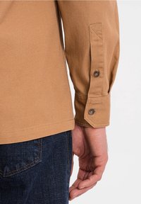 Brązowa koszula z fakturowanym wykończeniem, z mankietami zapinanymi na guziki i dopasowanym krojem, zestawiona z ciemnymi jeansami z detalem kieszeni.