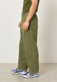 Zelené bavlněné kalhoty s uvolněným střihem, mají rovné nohavice, šité detaily a boční kapsy, doplněné o stříbrné a modré běžecké boty.