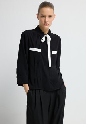Sort bluse med hvide accenter, plisseret design, krave og sløjfedetalje. Lange ærmer og løs pasform, parret med sorte, strukturerede bukser.