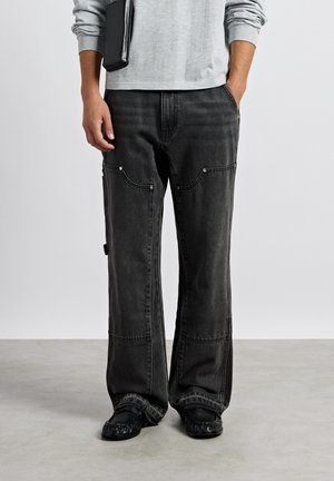 Homme portant un pantalon en denim noir ample avec des coutures visibles, des chaussures noires et une chemise à manches longues gris clair tenant une pochette noire.