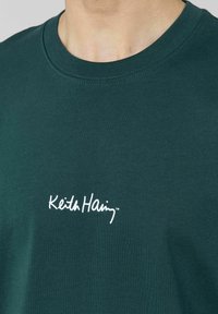 T-shirt en coton vert avec un col rond. Présente un logo blanc "Keith Haring" imprimé sur la poitrine. Texture lisse, design décontracté.