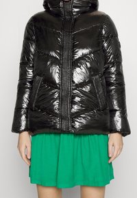 Chaqueta de plumas negra y brillante con capucha, con costuras verticales y dos bolsillos. Combinada con una falda verde, mostrando un estilo en capas.