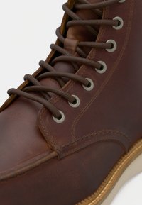 Botte en cuir brun avec des lacets ronds marron passés à travers des œillets en métal argenté et des coutures détaillées sur une semelle de couleur claire.
