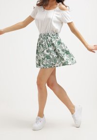 Kvinna i vit topp med kall axel och vit kjol med grönt blommönster, vita sneakers, står på ett ben med utsträckta armar.