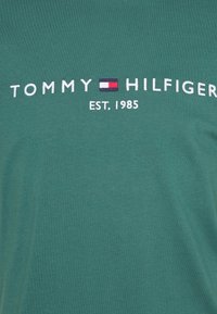 Vihreä puuvillainen t-paita, jossa on valkoisin kirjailuin teksti "TOMMY HILFIGER" ja pieni punaisen, valkoisen ja sinisen värinen logo. Perustettu 1985.