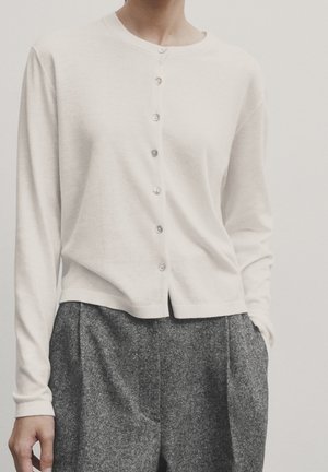 Personne portant un cardigan blanc à manches longues boutonné et un pantalon en laine gris taille haute, mains détendues sur les côtés, fond neutre.