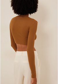 Kvinde med krøllet hår iført en tætsiddende, langærmet, cropped brun top og høj talje hvide bukser, stående mod en ensfarvet baggrund.