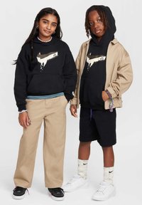 Hanorace negre cu design grafic, jachetă utilitară bej, pantaloni bej și pantaloni scurți negri. Adidași negri și albi. Îmbrăcăminte casual, texturată.