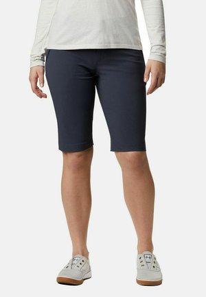 Person, die marineblaue knielange Shorts, ein weißes langärmeliges Hemd und weiße Slip-On-Schuhe vor einem einfarbigen Hintergrund trägt.