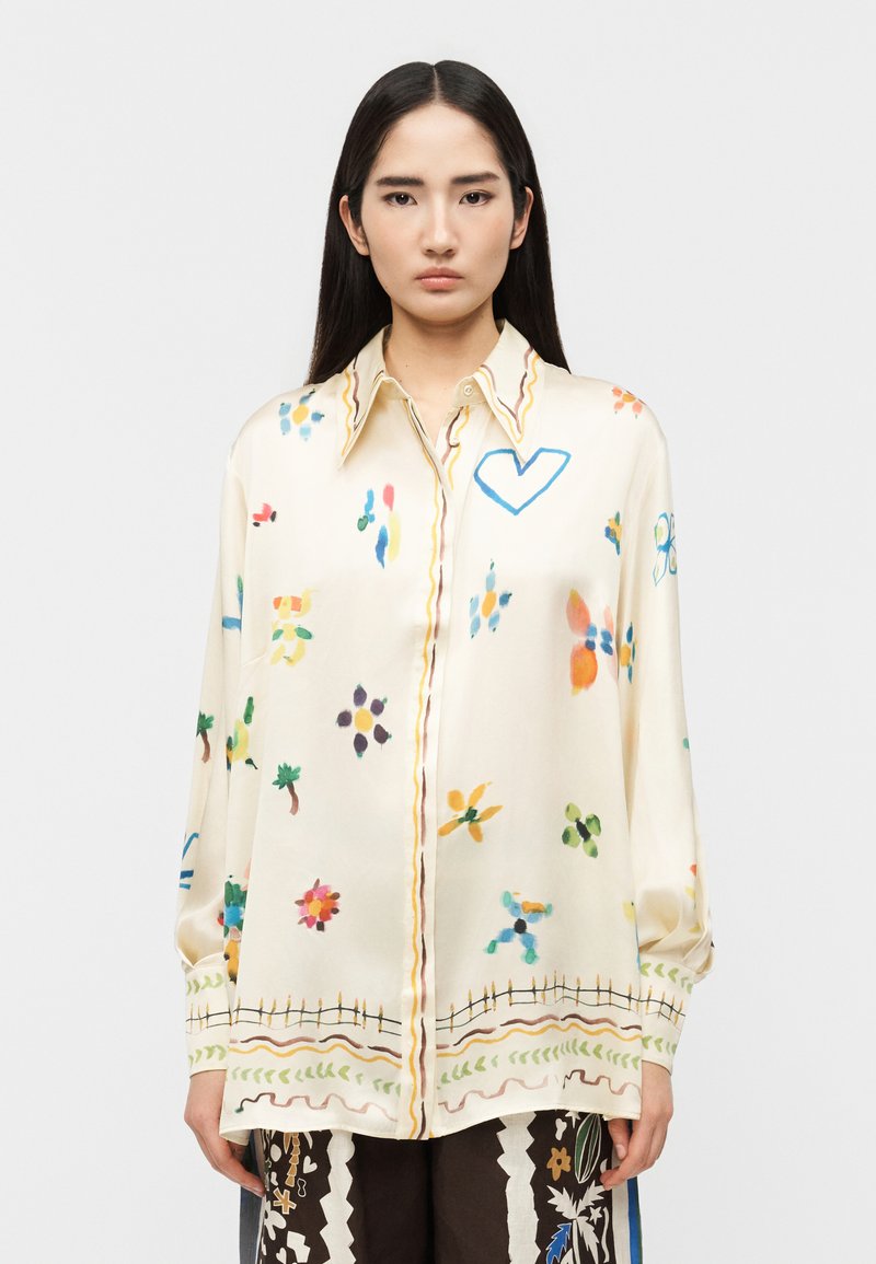 Γυναίκα με μακριά σκούρα μαλλιά φοράει υπερμεγέ θη κρεμ πουκάμισο με πολύχρωμα αφηρημένα λουλουδένια και καρδιές σχέδια, συνδυασμένο με μαύρο παντελόνι με σχέδια.