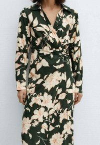 Robe portefeuille en tissu vert foncé avec de grands motifs floraux pêche, manches longues et col. Dotée d'un détail de taille nouée.