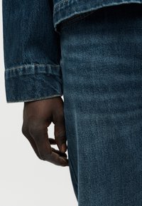 Μανίκι σκούρου μπλε τζιν μπουφάν και χέρι που ακουμπάει σε σκούρο μπλε τζιν παντελόνι. Το ύφασμα έχει μια απαλή υφή με διακριτικές λεπτομέρειες ραφής σε αντιθετικό χρώμα.