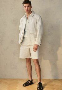 Licht beige jas met twee voorke pockets, over een bijpassend top met korte mouwen en shorts. Zwarte sandalen met bandjes maken de outfit compleet.