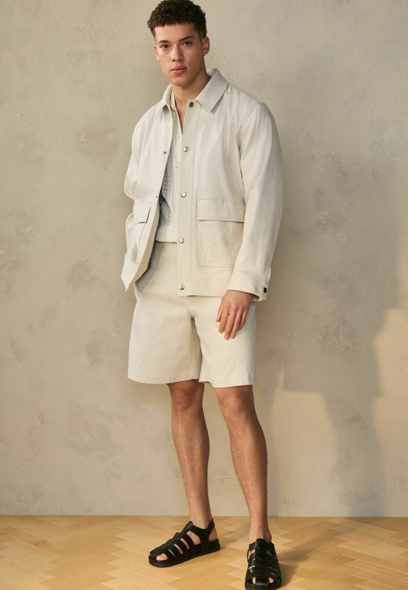 Licht beige jas met twee voorke pockets, over een bijpassend top met korte mouwen en shorts. Zwarte sandalen met bandjes maken de outfit compleet.