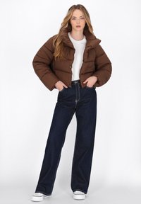 Giacca puffer marrone con colletto alto, chiusura a bottone e texture trapuntata, abbinata a jeans a gamba larga blu scuro e sneakers bianche.