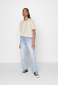 T-shirt beige a maniche corte con logo sul lato sinistro del petto, abbinato a jeans a gamba larga blu chiaro e sneakers nere alte.