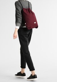 Bordová taška na stažení s jemnou texturou. Nositelná na zádech, spárovaná s černými kalhotami a botami. Jednoduchý design, malé logo viditelné.