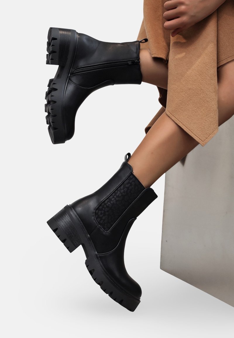 Des bottines noires présentent une tige en cuir lisse, des panneaux latéraux texturés, une semelle crantée épaisse et une fermeture à zip à l'arrière. Les accents incluent une languette de traction.