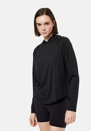 Jeune femme portant un haut à capuche noir à manches longues et un short noir moulant, se tenant devant un fond clair uni.