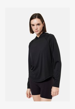 Jeune femme portant un haut à capuche noir à manches longues et un short noir moulant, se tenant devant un fond clair uni.