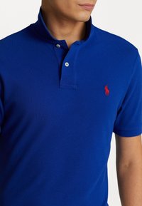 Blå pikétröja i bomullsmaterial med en strukturerad väv, med en tvåknappsknäppning och en röd broderad logotyp på vänster bröst.