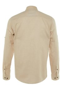 Chemise beige à manches longues avec poignets boutonnés, vue de dos sur fond blanc uni.