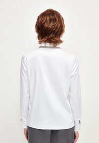 Persona dai capelli rossi che indossa una camicia bianca a maniche lunghe con un colletto grigio, in piedi di spalle contro uno sfondo chiaro e semplice.