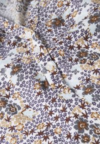 Tissu à motif floral bleu clair, marron et gris avec de petites fleurs et feuilles. Doté de boutons blancs sur le devant.