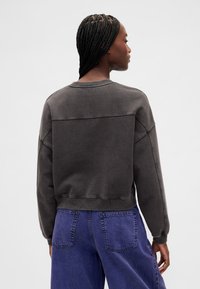 Tmavě šedá mikina s kulatým výstřihem a zkráceným střihem, s nízkými rameny a hladkou texturou. Kombinována s modrými džíny s vysokým pasem.